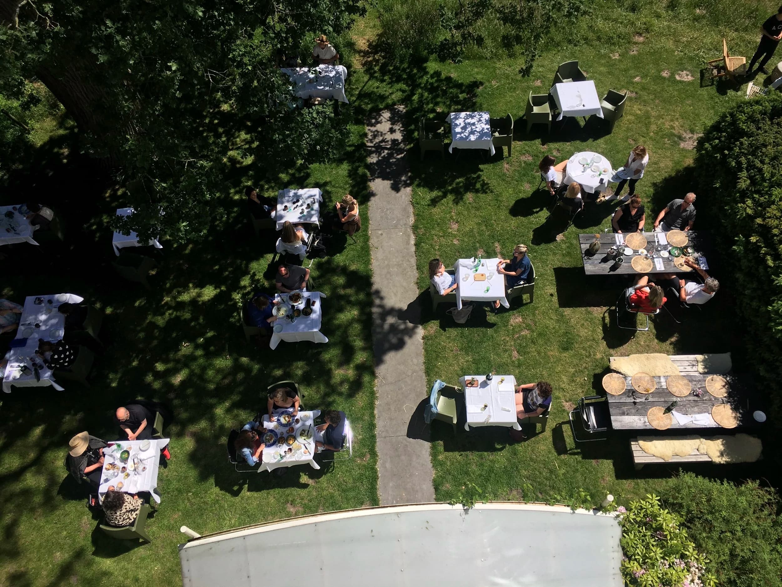 Buitenevenement met mensen die aan tafels zitten in een tuin, gezien vanaf boven.