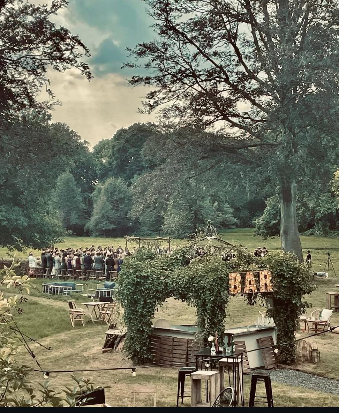 Een buitenevenement in een park met een bar, omringd door bomen, en een groot groep mensen onder een houten structuur.