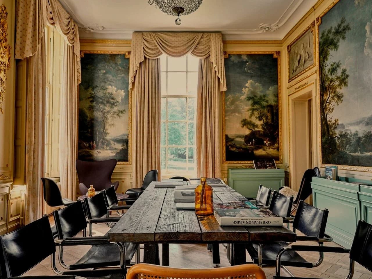 Een vergaderruimte met een grote houten tafel, zwarte stoelen, muurkunst met landschapstafereel, grote ramen met gordijnen, een groene kast, en decoratieve plafonddetails.