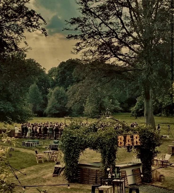 Een buitenfeest met mensen die samen zingen of een optreden doen, omgeven door bomen en een gezellige bar met groen decor.