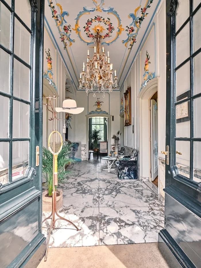 Elegante entreehal met marmeren vloer, kroonluchter, wit plafond met kleurrijk decor, en vintage meubels en decoraties.