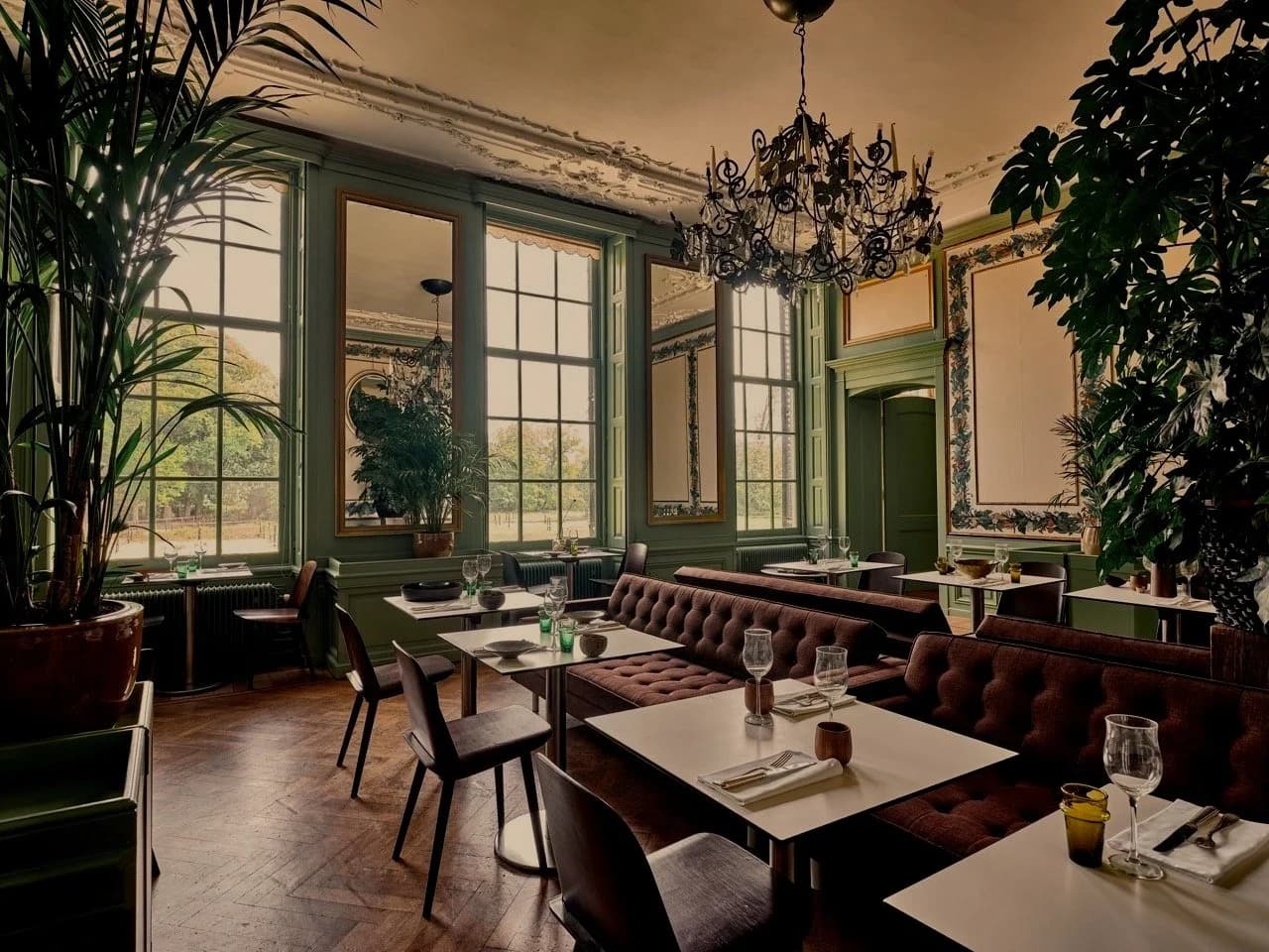 Elegante restaurant met grote ramen, chandelier, groene muren, planten, en gedekte tafels.