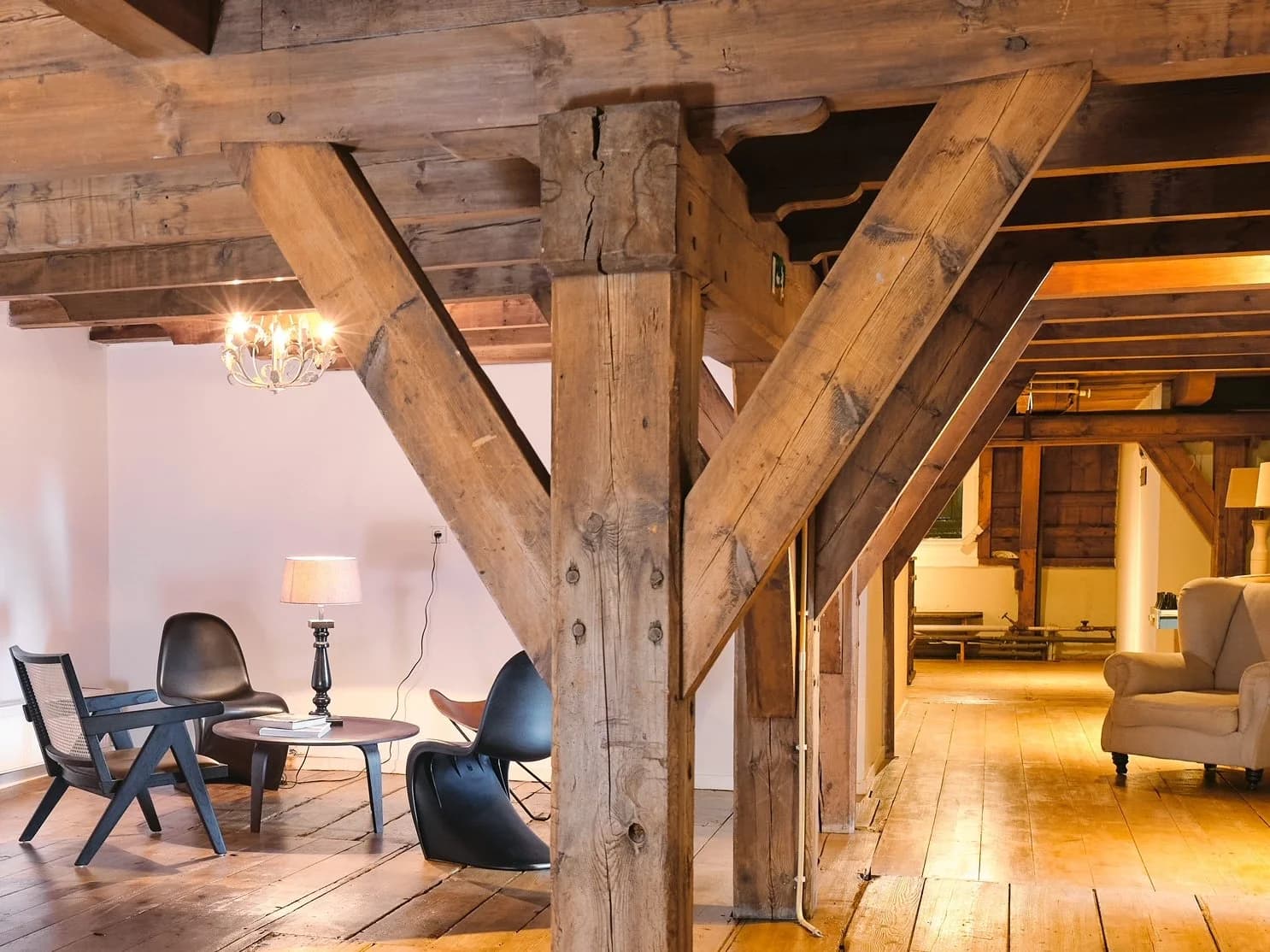 Interieur van een kamer met houten vloer en houten plafond, met meubels zoals een zwarte stoel, een bijzettafel met boeken en een tafellamp, en een beige bank. In het midden een grote houten steunbalk en truss.
