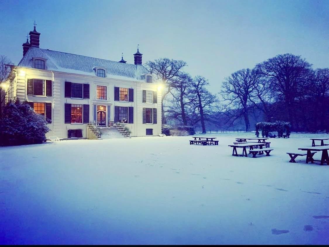 Een wit herenhuis met zwarte luiken en een schuine dak, verlicht door buitenverlichting, omgeven door een besneeuwd landschap en enkele blikken tafels en banken in de sneeuw, met bomen op de achtergrond in de schemering.