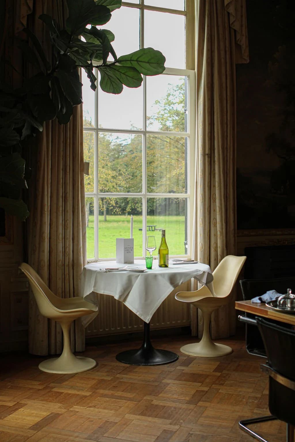 Een etentafel met een wit tafelkleed, glazen, wijnfles, en een menu, voor een groot raam met uitzicht op een groene tuin in een klassieke interieurruimte.